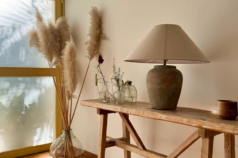 Wooden console table with a ceramic lamp, glass vases with pampas grass, and decorative items near a sunlit window