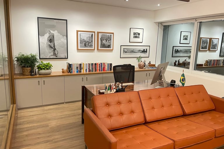 Modern office interior with an orange leather couch, wooden storage cabinets, framed artwork on walls, and glass partitions separating workspaces.