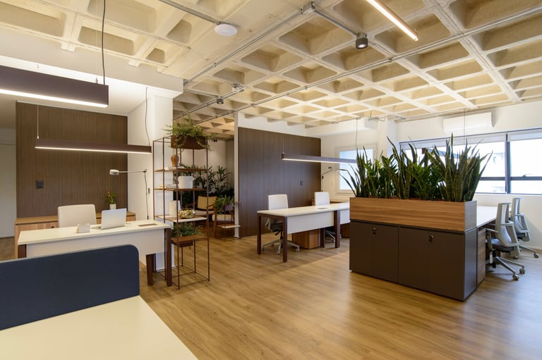 Modern office space with wooden flooring, glass-enclosed meeting rooms, white desks, and potted plants throughout